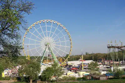 Ein großes Riesenrad dominiert die Mitte eines festlichen Jahrmarkts, darunter befinden sich verschiedene farbenfrohe Fahrgeschäfte und Buden. Bäume rahmen den Vordergrund ein und unter einem klaren blauen Himmel herrscht reges Treiben.