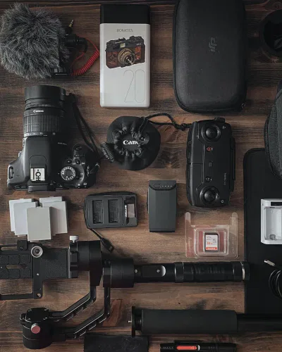 Eine flache Kameraausrüstung auf einer hölzernen Unterlage, perfekt für die Aufnahme von Frankfurter Veranstaltungen oder die Erstellung kreativer Video-Ideen, mit einer Canon DSLR, Objektiven, Speicherkarten, Batterien, Gimbal-Stabilisator, Mikrofon mit Windschutz und mehr.