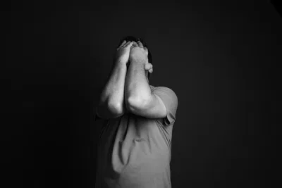 Eine Person, die ein T-Shirt trägt, steht vor einem dunklen Hintergrund und bedeckt ihr Gesicht mit beiden Händen und angewinkelten Ellbogen, was auf organisierten Stress oder Bedrängnis hindeutet - vielleicht spiegelt es Abschiede oder emotionale Belastungen wider, denen Angehörige ausgesetzt sind. Das Bild ist in schwarz-weiß gehalten.