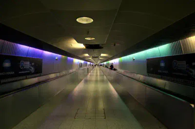 Ein langer, leerer Flughafenkorridor mit einem Laufband, beleuchtet von Decken- und mehrfarbigen Seitenlichtern am Flughafen Frankfurt. An der Decke befinden sich mehrere Scheinwerfer, und an den Wänden hängen Werbeanzeigen. Das Laufband verschwindet in der Ferne und suggeriert Tiefe und Perspektive.