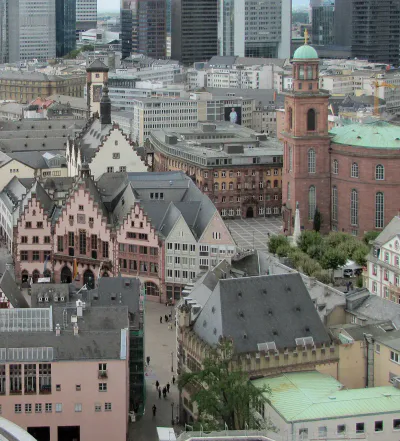 Luftaufnahme von Frankfurt, Deutschland, mit einer Mischung aus historischer und moderner Architektur. Zu den markanten Gebäuden zählen der mittelalterliche Römer in der Altstadt mit seinen Stufengiebeln und die Paulskirche aus rotem Sandstein inmitten der umliegenden Wolkenkratzer und der Stadtlandschaft.