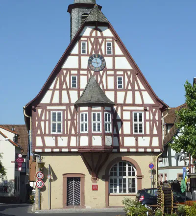 Ein Fachwerkhaus mit steilem, dreieckigem Dach und einem kleinen Türmchen mit einer Uhr steht im hellen Tageslicht von Bergen-Enkheim. Das Gebäude hat ein beiges Erdgeschoss mit einem gewölbten Eingang. Rundherum sind saubere Straßen, Verkehrsschilder und Grünflächen.