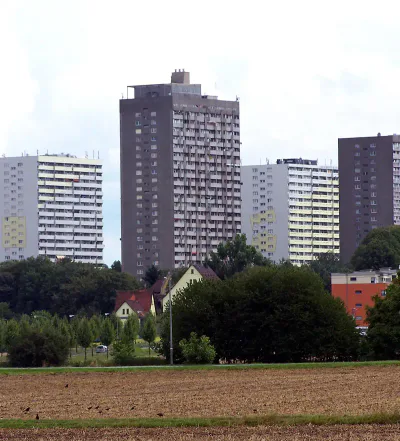 frankfurt-stadtteile-frankfurter-berg Eine Landschaft mit mehreren hohen Wohnhäusern hinter einer Ansammlung kleinerer Häuser, Bäumen und Grünflächen im Frankfurter Berg. Im Vordergrund ist ein offenes Feld mit Gras- und Erdflecken zu sehen.