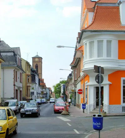 Eine malerische europäische Straße in Heddernheim, gesäumt von farbenfrohen Gebäuden und an beiden Seiten geparkten Autos. Ein leuchtend orange-weißes Gebäude ziert die Ecke, während im Hintergrund unter einem teilweise bewölkten Himmel eine Kirche mit einem Glockenturm hervorsticht.