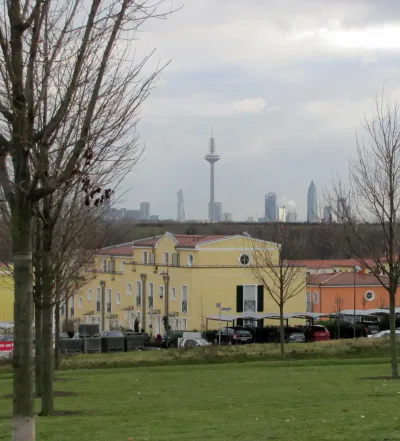 Ein Park mit Bäumen und grünem Gras im Vordergrund, Gebäude mit gelben und orangefarbenen Fassaden im Mittelgrund und eine Stadtsilhouette mit modernen Hochhäusern und einem markanten Funkturm im Hintergrund unter einem wolkigen Himmel, die an die malerische Aussicht von Kalbach-Riedberg erinnert.