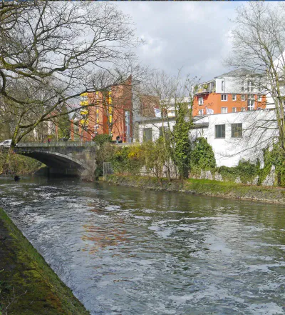Ein ruhiger Fluss fließt unter einer kleinen Brücke in Rödelheim hindurch, einem Waldgebiet neben Wohnhäusern. Entlaubte Bäume säumen die Ufer, während im Hintergrund Backstein- und weiße Gebäude zu sehen sind. Die Szene wird vom hellen Tageslicht unter einem teilweise bewölkten Himmel erhellt.