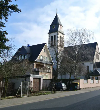 frankfurt-stadtteile-schwanheim In einer ruhigen Straße in Schwanheim steht ein beiges, zweistöckiges Haus mit hölzernen Fensterläden und einem Balkon. Daneben steht ein hohes Kirchengebäude mit einem Glockenturm. Beide Gebäude sind von kahlen Bäumen umgeben und neben dem Haus stehen an einer Steinmauer zwei geparkte Autos.