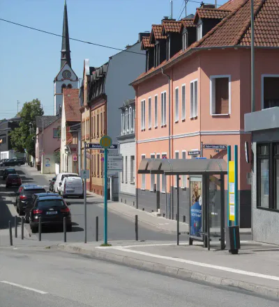 Eine ruhige Straße in der europäischen Stadt Sossenheim ist geprägt von mehrstöckigen Gebäuden in Pastellfarben. Die Straße bietet eine Bushaltestelle mit Unterstand auf der rechten Seite, geparkte Autos entlang der Straße und im Hintergrund eine Kirche mit einem Glockenturm unter einem klaren blauen Himmel.
