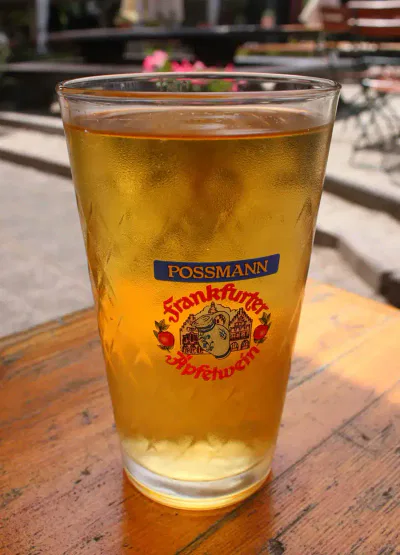 Ein Glas mit einem gelben Getränk, wahrscheinlich Apfelwein, mit dem Logo „Possmann Frankfurter Apfelwein“ auf der Vorderseite. Das Glas steht auf einem Holztisch im Freien, mit verschwommenen Hintergrundelementen wie Stühlen und Pflanzen.