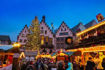 Ein festlicher Weihnachtsmarkt am Abend mit einem großen, beleuchteten Weihnachtsbaum vor historischen Gebäuden. Die Marktstände sind mit Lichterketten geschmückt und eine Menschenmenge genießt die festliche Atmosphäre bei diesem lebhaften Fest.