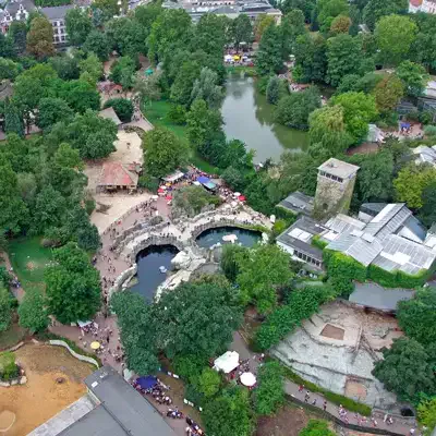 Luftaufnahme eines großen Zoos, umgeben von üppigen grünen Bäumen. Zu sehen sind verschiedene Gehege, ein zentraler Teich, mit Besuchern gefüllte Spazierwege und mehrere Gebäude. Die Szene ist voller Aktivität und natürlicher Sehenswürdigkeiten.