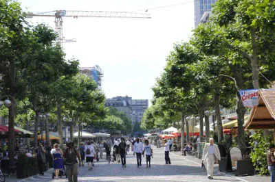 Eine belebte Fußgängerzone, gesäumt von Bäumen und Caféterrassen auf beiden Seiten, strahlt eine festliche Atmosphäre aus. Die Menschen gehen spazieren, sitzen und gehen verschiedenen Aktivitäten nach. Im Hintergrund sind ein Kran und eine Baustelle zu sehen, was auf eine laufende Entwicklung in der Gegend schließen lässt.