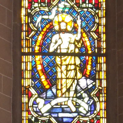 Ein Buntglasfenster in der Alten Nikolaikirche zeigt eine religiöse Figur, wahrscheinlich Jesus, in einem Gewand mit einem Heiligenschein hinter dem Kopf, der eine Hand erhebt. Das Glas ist in leuchtenden Farben wie Blau, Rot, Gelb und Grün gehalten und mit komplizierten dekorativen Mustern geschmückt.