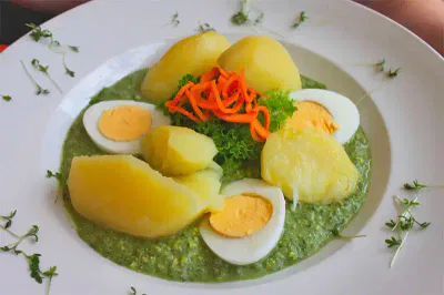 Ein Teller mit gekochten gelben Kartoffeln, geschnittenen hartgekochten Eiern und einer leuchtend grünen Soße. Das Gericht ist mit krauser Petersilie und dünnen Streifen roter Paprika garniert. Dünne grüne Garnituren sind um den Rand des weißen Tellers verstreut.