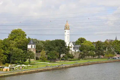 Ein malerischer Blick auf das Flussufer mit einem hohen, weißen Turm mit goldenem Dach, umgeben von üppigen grünen Bäumen und Gebäuden. Die Menschen entspannen auf dem Gras und an Tischen im Freien entlang des Flussufers und genießen die festliche Atmosphäre, während sich über ihnen Stromleitungen durch den Himmel ziehen.