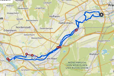 Eine detaillierte Karte zeigt eine Route von Frankfurt, gekennzeichnet mit einem „A“, durch mehrere Städte, darunter Hofheim, Hattersheim und Raunheim. Die Route folgt dem Main und führt durch Grüngebiete wie Mönchbruch von Mörfelden und Rüsselsheim.