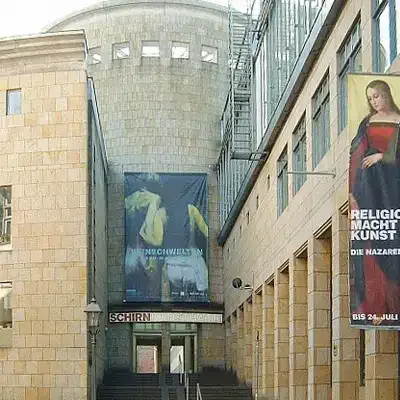 Ein Steingebäude mit Bannern am Eingang. Ein Banner zeigt ein Kunstwerk mit gelben und schwarzen Elementen, während das andere ein Gemälde einer religiösen Figur mit deutschem Text zeigt. Das Gebäude, das oft als eine der lokalen Sehenswürdigkeiten angesehen wird, verfügt über Stufen, die zum Eingang hinaufführen.