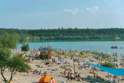 Eine überfüllte Strandszene mit zahlreichen Menschen, die sich sonnen, schwimmen und entspannen. Am Sandstrand sind verschiedene Sonnenschirme und Zelte verteilt. Das klare blaue Seewasser ist von grünen Bäumen und einem dichten Wald im Hintergrund umgeben.