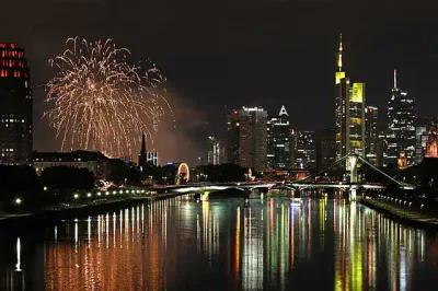Eine nächtliche Stadtlandschaft mit einem Fluss, dessen Gebäude und eine Brücke bunte Lichter reflektieren. Auf der linken Seite explodieren Feuerwerke am Himmel, fügen lebendige Farben hinzu und erhellen die festliche Szene.