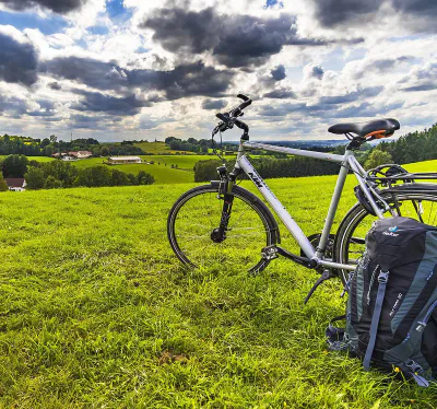 Auf einer grünen Wiese steht ein silbernes Fahrrad mit schwarzem Sattel und Lenker. Daneben liegt ein schwarzer Rucksack. Im Hintergrund sind sanfte Hügel, Bäume und bewölkter Himmel zu sehen. Die Szene vermittelt ein Gefühl von Ruhe und Abenteuer.