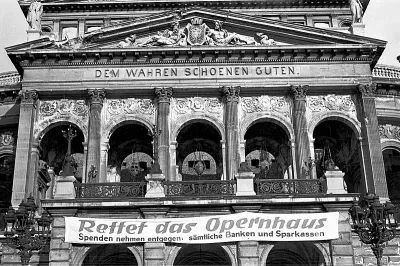 Eine historische Opernhausfassade mit raffinierter Architektur und einem Banner mit der Aufschrift „Rettet das Opernhaus“ und darunter einem zusätzlichen Text zu Spenden. Über den Bögen ist der Satz „DEM WAHREN SCHOENEN GUTEN“ eingraviert. Dieses ikonische Gebäude ist kein anderes als die Alte Oper.