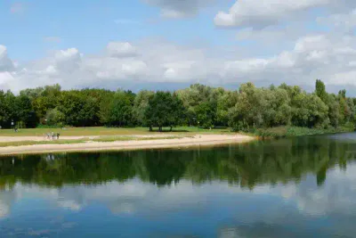 Eine ruhige Szene am Seeufer mit ruhigem Wasser, das einen teilweise bewölkten Himmel und dichte grüne Bäume widerspiegelt. Am grasbewachsenen Ufer gehen ein paar Leute spazieren und genießen die Ruhe. Das Ufer besteht aus einer Mischung aus Sand und Gras und schafft eine friedliche Naturlandschaft.