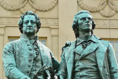 Eine Bronzestatue zeigt zwei historische Figuren, die Seite an Seite vor einem neoklassizistischen Gebäude stehen. Beide tragen Mäntel aus dem 18. Jahrhundert und halten Lorbeerkränze. Einer von ihnen hat seinen Arm um Goethes Schulter gelegt. Die beiden stehen vor einem Hintergrund, der mit dekorativen architektonischen Details geschmückt ist.