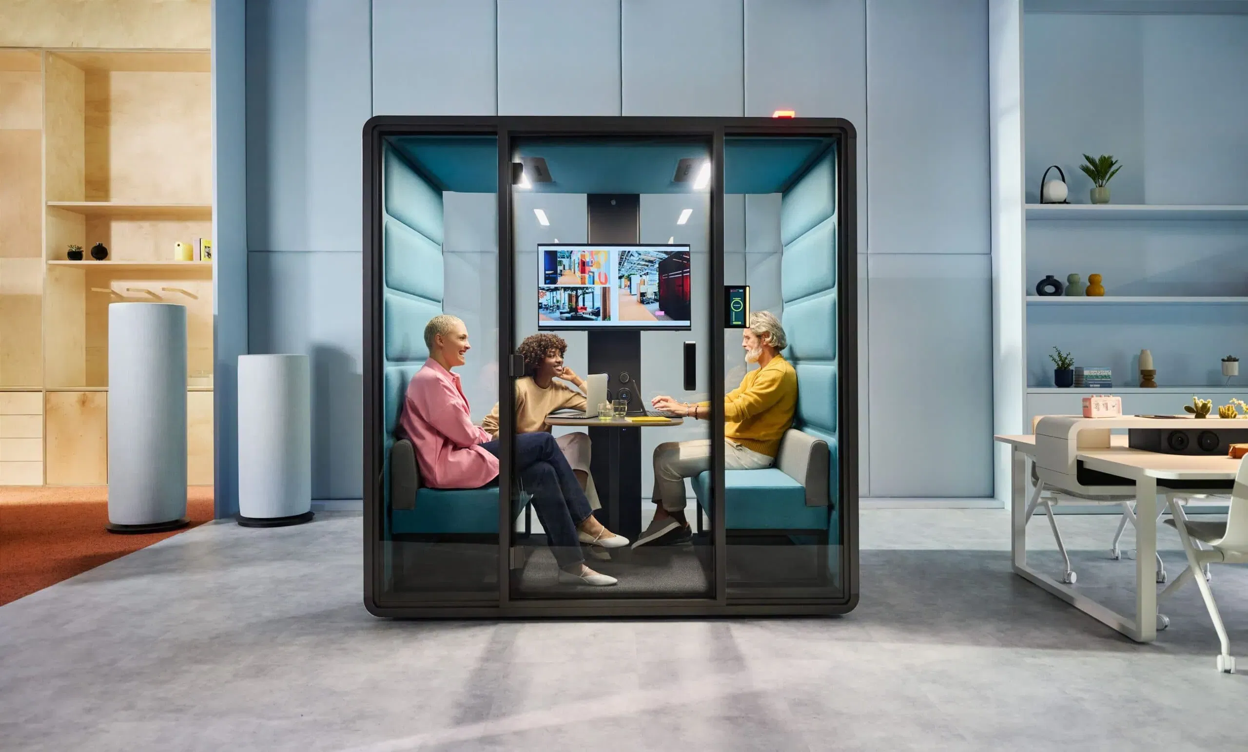 Drei Personen sitzen in einem modernen, verglasten Konferenzraum mit schalldichten Wänden und unterhalten sich. Der Raum ist mit blauen Kissen ausgestattet und verfügt über einen digitalen Bildschirm, auf dem Diagramme angezeigt werden. Der umliegende Büroraum verfügt über Regale und einen Schreibtisch mit Dekorationsgegenständen.