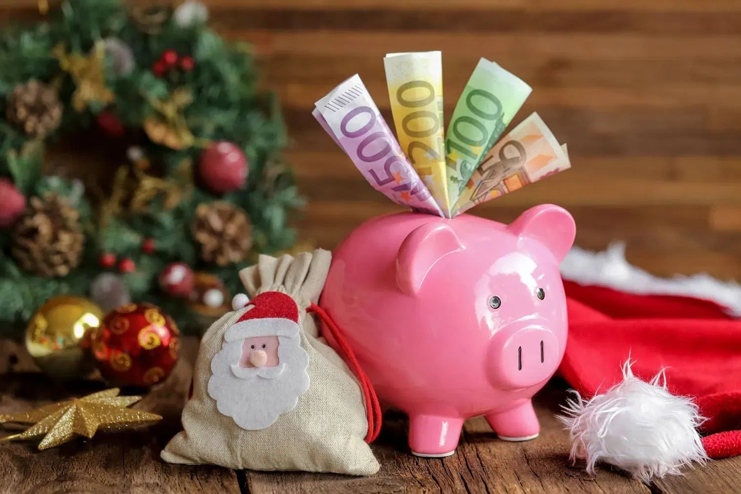 Ein rosa Sparschwein ist randvoll mit Euro-Scheinen, daneben steht ein kleiner Sack mit dem Weihnachtsmann, der auf die Faszination von El Gordo anspielt. Im Hintergrund zieren ein festlicher Weihnachtskranz und dekorative Ornamente die Holzoberfläche und wecken Träume von einem unerwarteten Weihnachtssegen.