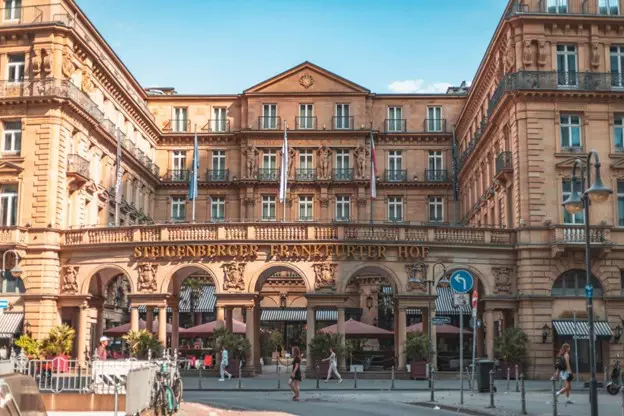 Das historische Grandhotel mit kunstvoller Architektur namens „Steigenberger Frankfurter Hof“ verkörpert die Eleganz Frankfurts. Seine beigefarbene Fassade, mehrere Stockwerke und der gewölbte Eingang heißen Passanten willkommen. Sitzbereiche im Freien mit Sonnenschirmen laden zum Entspannen unter dem klaren blauen Himmel ein.