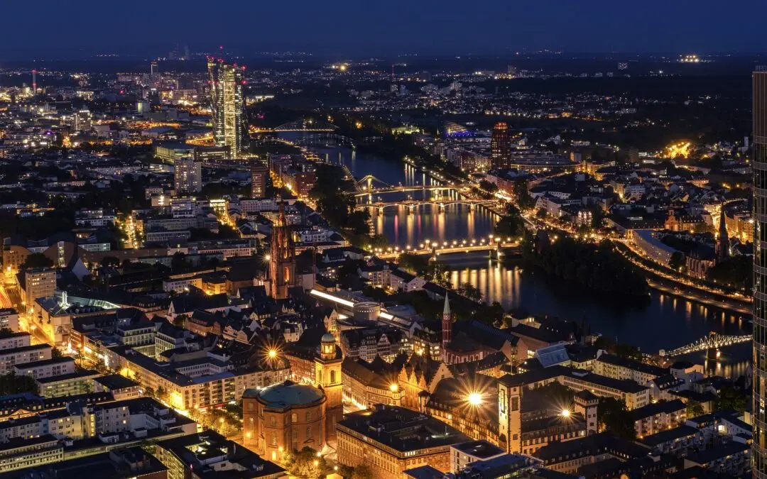 Ein nächtlicher Panoramablick auf die Mainmetropole, beleuchtet mit hellen Lichtern. Das Bild zeigt einen Fluss, der durch die Stadt fließt, mehrere Brücken, die beide Seiten verbinden, sowie markante Gebäude und Wolkenkratzer. Der Himmel ist dunkel und die Lichter erzeugen eine lebendige Stadtlandschaft, die an virtuelle Glücksspiele erinnert.