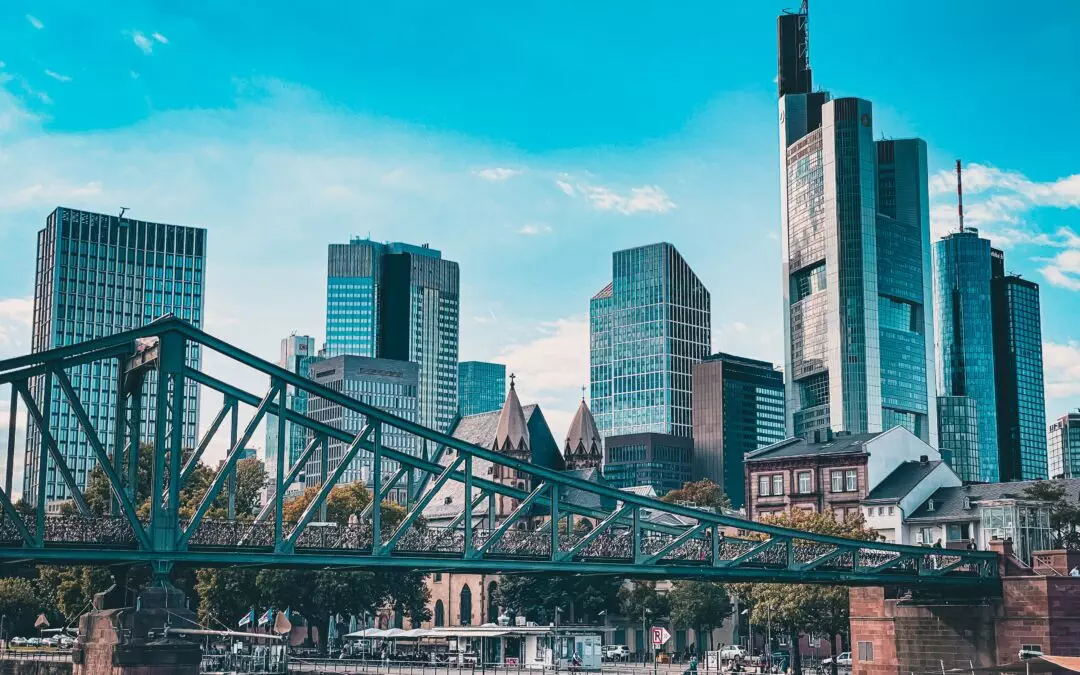 Ein malerischer Blick auf die Skyline von Frankfurt mit hohen, modernen Wolkenkratzern vor einem strahlend blauen Himmel. Im Vordergrund ist eine grüne Metallbrücke über einen Fluss zu sehen, mit Gebäuden und grünen Bäumen entlang des Flussufers. Das Wasser spiegelt die Strukturen und den Himmel und bietet zahlreiche Möglichkeiten zum Fotografieren.