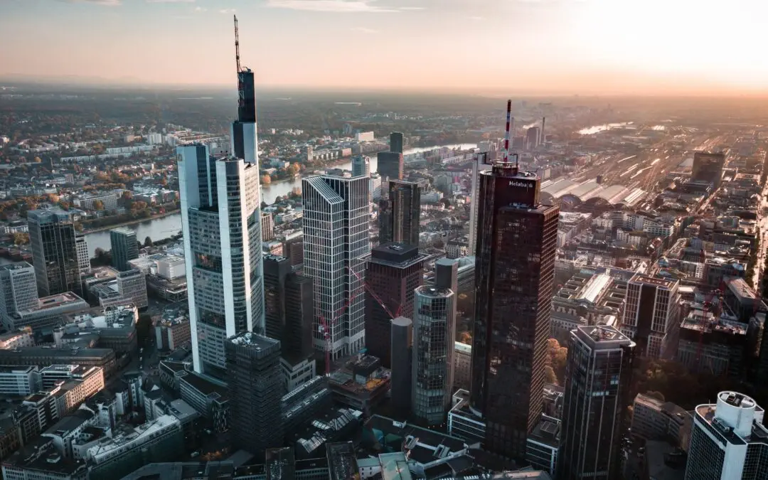 Luftaufnahme der Frankfurter Skyline bei Sonnenuntergang. Die Skyline wird von modernen Wolkenkratzern, darunter dem Commerzbank Tower, und anderen Hochhäusern dominiert. Man kann den Main sehen, der sich durch die Stadt schlängelt, und in der Ferne erstrecken sich weitläufige Stadtgebiete dieser pulsierenden Mainstadt.