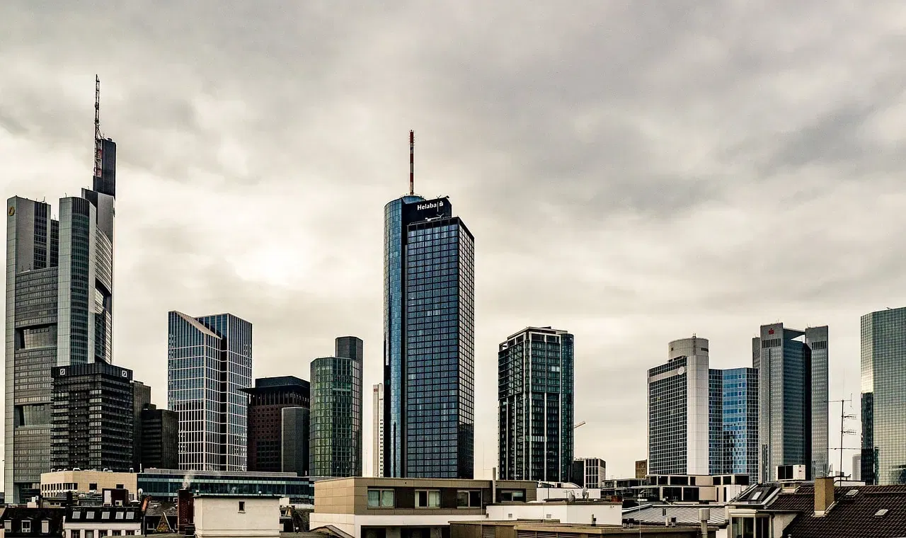 Eine Skyline mit modernen Hochhäusern und Wolkenkratzern unter einem bewölkten Himmel in Frankfurt am Main. Die Architektur besteht hauptsächlich aus Glas- und Stahlkonstruktionen, die eine zeitgenössische, urbane Atmosphäre schaffen. Im Vordergrund sind Dächer und kleinere Gebäude zu sehen, perfekt für diejenigen, die ihr perfektes Zuhause suchen.