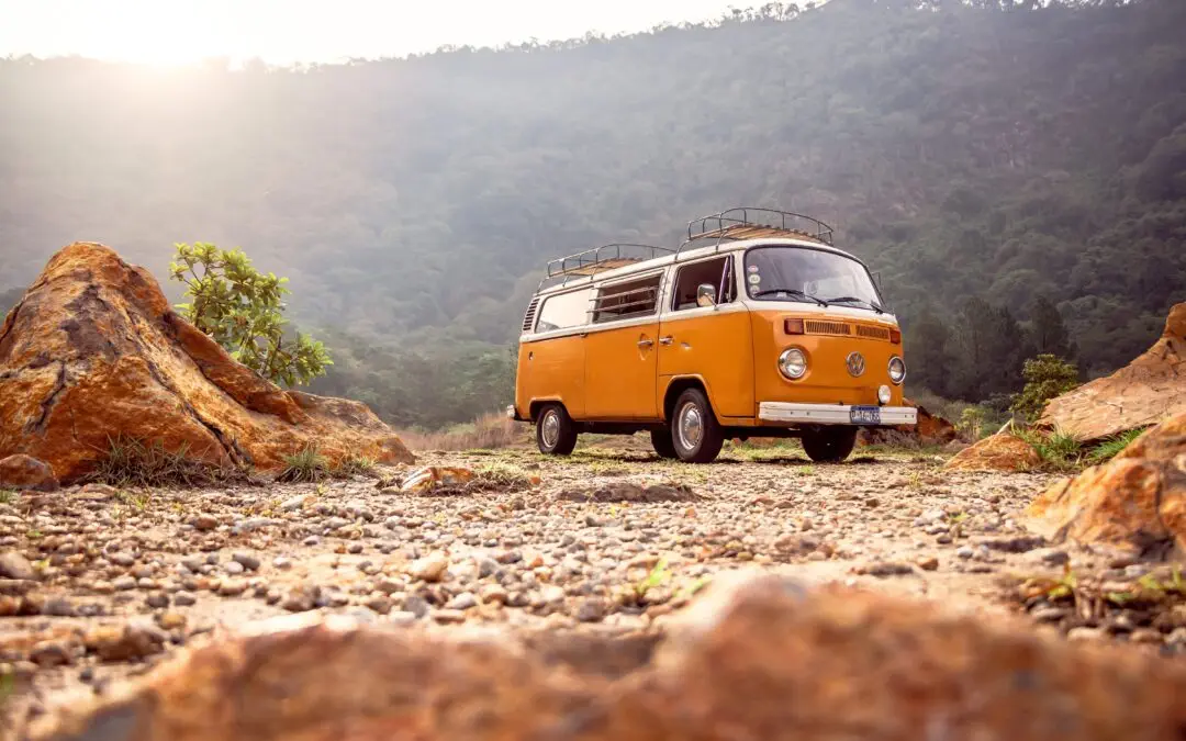 Ein orangefarbener Oldtimer-Van, perfekt für eine Hochzeit, steht auf felsigem Gelände mit grünen Hügeln und einem teilweise bewölkten Himmel im Hintergrund. Die Sonne scheint durch die Wolken und wirft ein warmes Licht auf die Szene, was den Charme dieses Fotobusses unterstreicht, der ideal ist, um bleibende Erinnerungen zu schaffen.