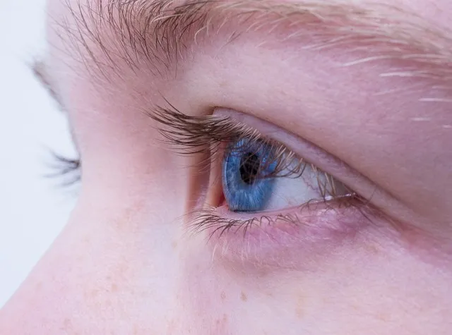 Seitliche Nahaufnahme des Gesichts einer Person, wobei der Schwerpunkt auf einem blauen Auge mit langen Wimpern und heller Haut liegt. Augenbrauen und Hauttextur sind sichtbar und heben die für das Sehen wichtigen Details hervor. Der Hintergrund ist unscharf und von heller Farbe.