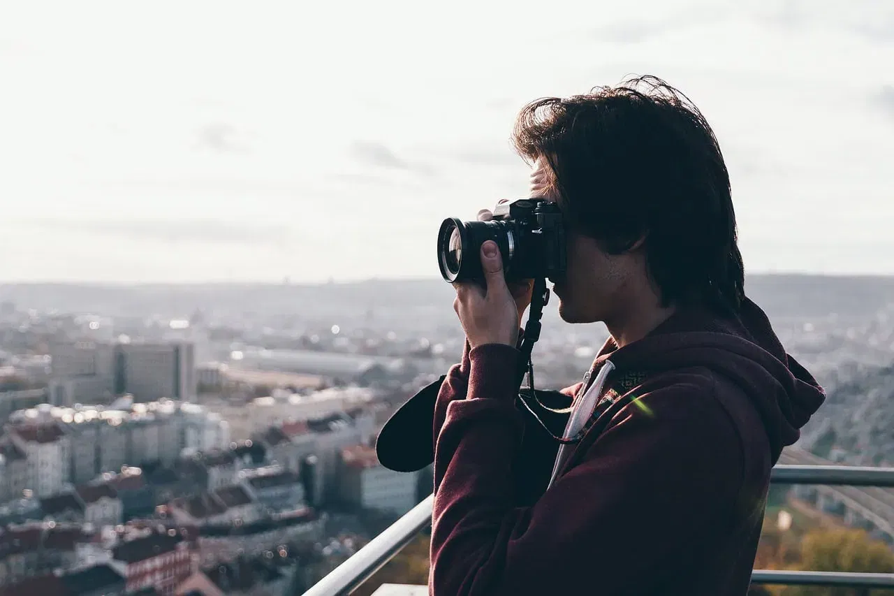 Eine Person mit schulterlangem Haar fotografiert mit einer Kamera von einem hohen Aussichtspunkt aus, von wo aus sie die weitläufige Stadtlandschaft Frankfurts unter einem bewölkten Himmel überblickt. Sie trägt einen kastanienbraunen Kapuzenpullover und hält die Kamera ans Auge. So macht sie Instagram-würdige Fotos.