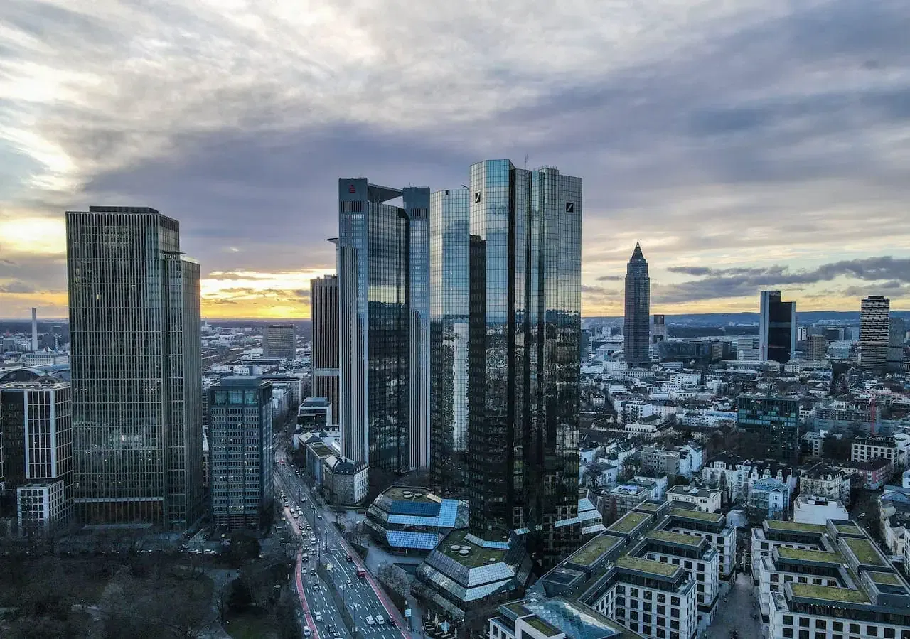Luftaufnahme der Frankfurter Skyline mit hohen, modernen Wolkenkratzern mit Glasfassaden. Im Hintergrund ist der Sonnenuntergang zu sehen, am Himmel ziehen Wolken auf. Im Vordergrund sind belebte Straßen und kleinere Gebäude zu sehen, darunter eine bepflanzte Dachterrasse in der Nähe der Krypto-Regulierungsbehörde.