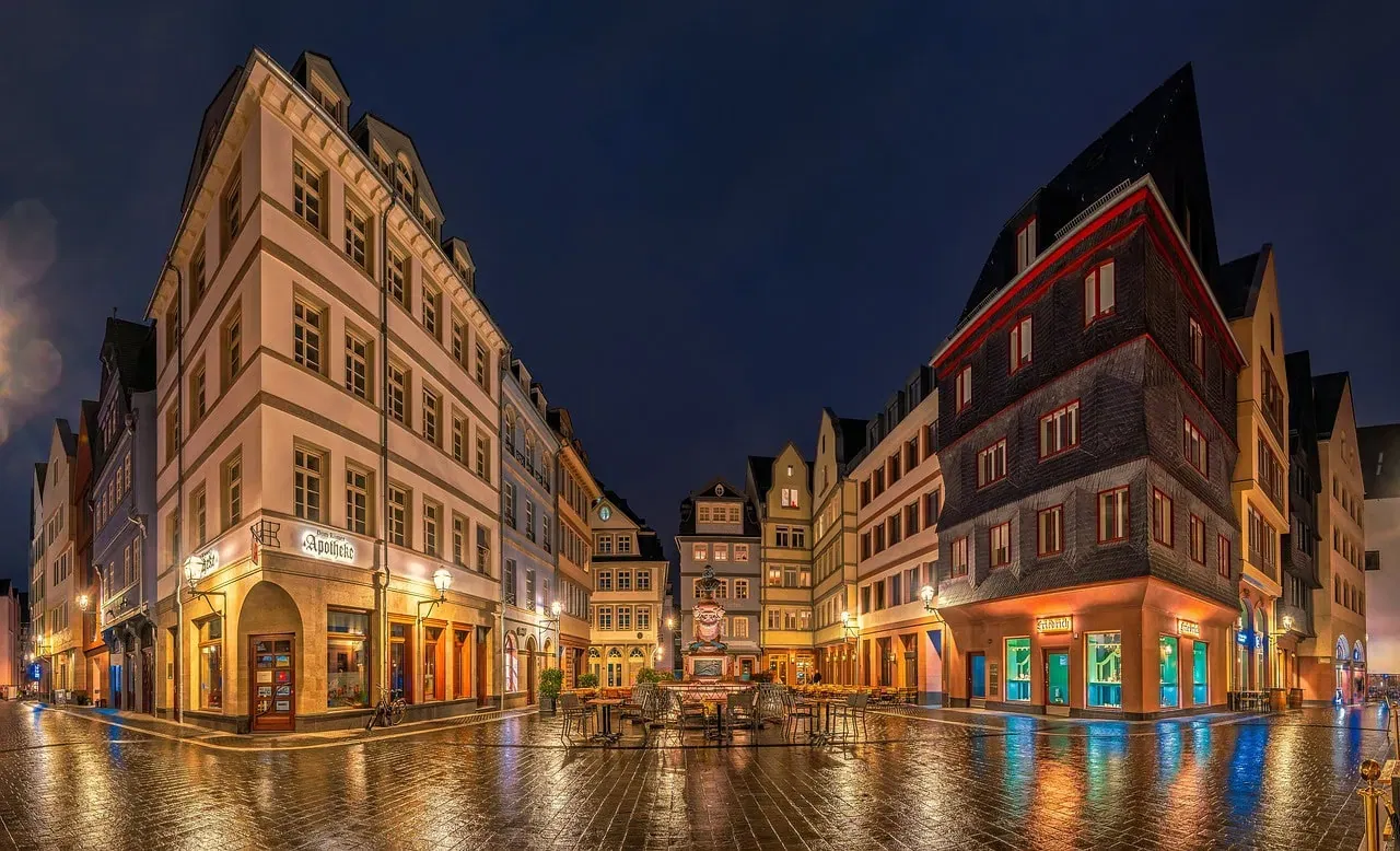 Ein wunderschön beleuchteter Stadtplatz bei Nacht mit traditioneller europäischer Architektur, der Frankfurts nächtlichen Charme ausstrahlt. Die Szene zeigt eine Kopfsteinpflasterstraße, historische Gebäude mit warmer Beleuchtung, einen kleinen Brunnen in der Mitte und trendige Bars an den Ecken.