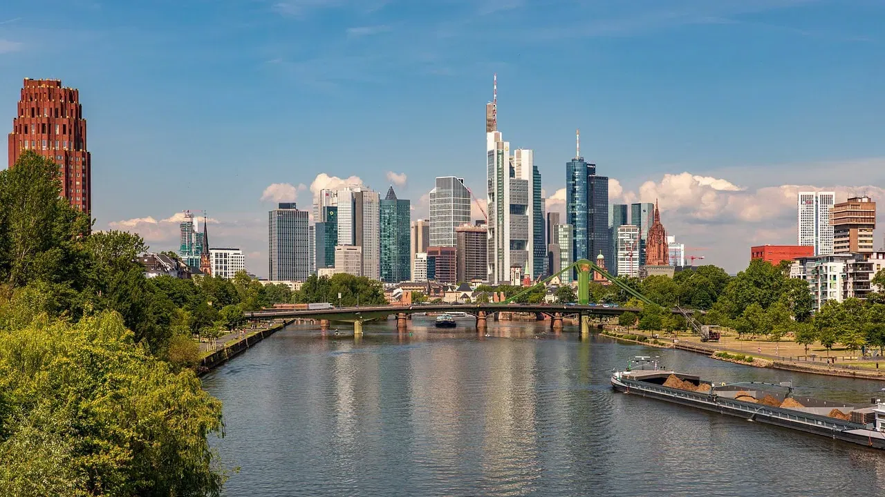 Klare Sicht auf die Skyline von Frankfurt mit modernen Wolkenkratzern unter blauem Himmel. Im Vordergrund fließt der Main, in dem sich die Gebäude spiegeln. Grüne Bäume säumen das Flussufer, eine Brücke überspannt den Fluss, auf dem Boote zu sehen sind – eine ideale Kulisse für die Diskussion steigender Preise.