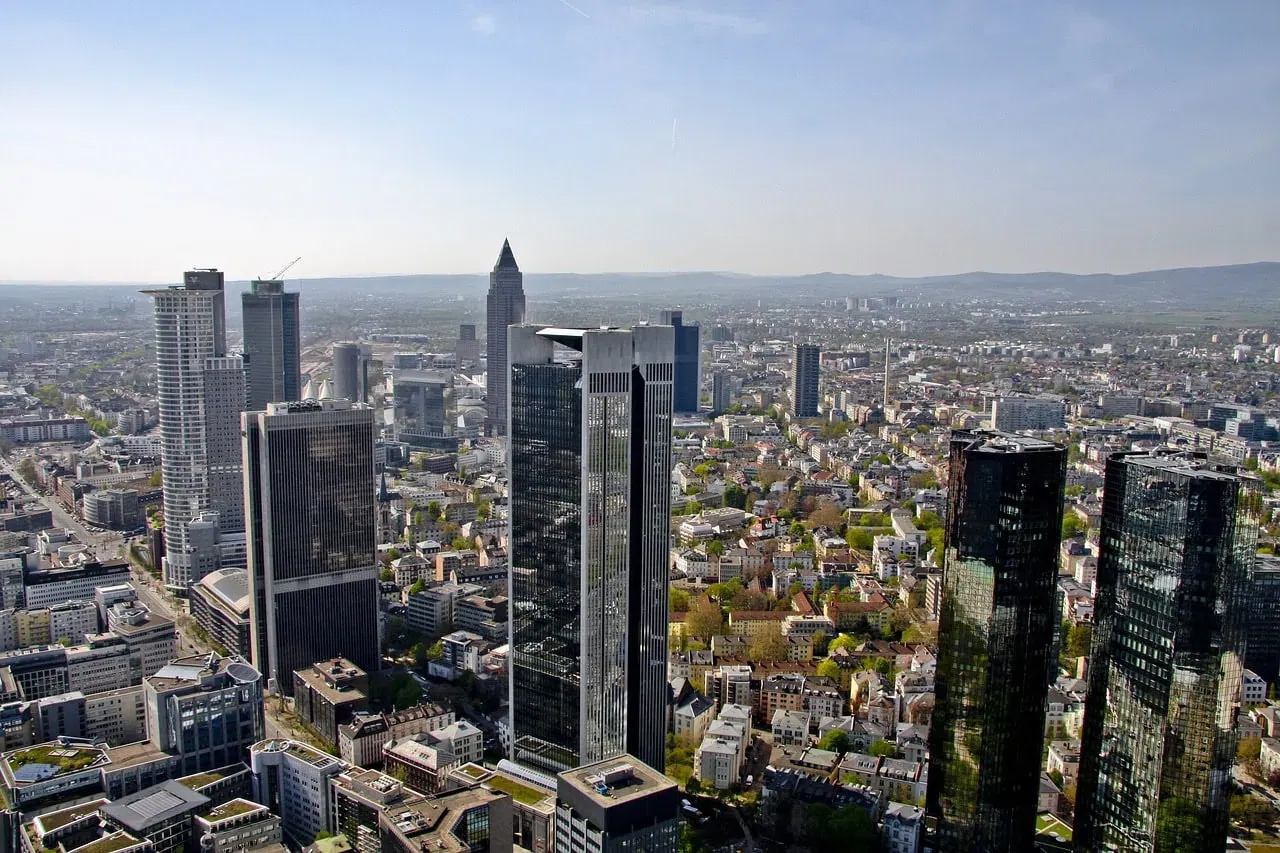 Luftaufnahme einer geschäftigen Skyline in Deutschland, die moderne Wolkenkratzer neben Wohn- und Geschäftsgebäuden zeigt. Die ausgedehnte Landschaft bietet üppige Grünflächen und ferne Hügel unter einem klaren blauen Himmel, ganz wie die lebendigen Welten von Online-Spielen.