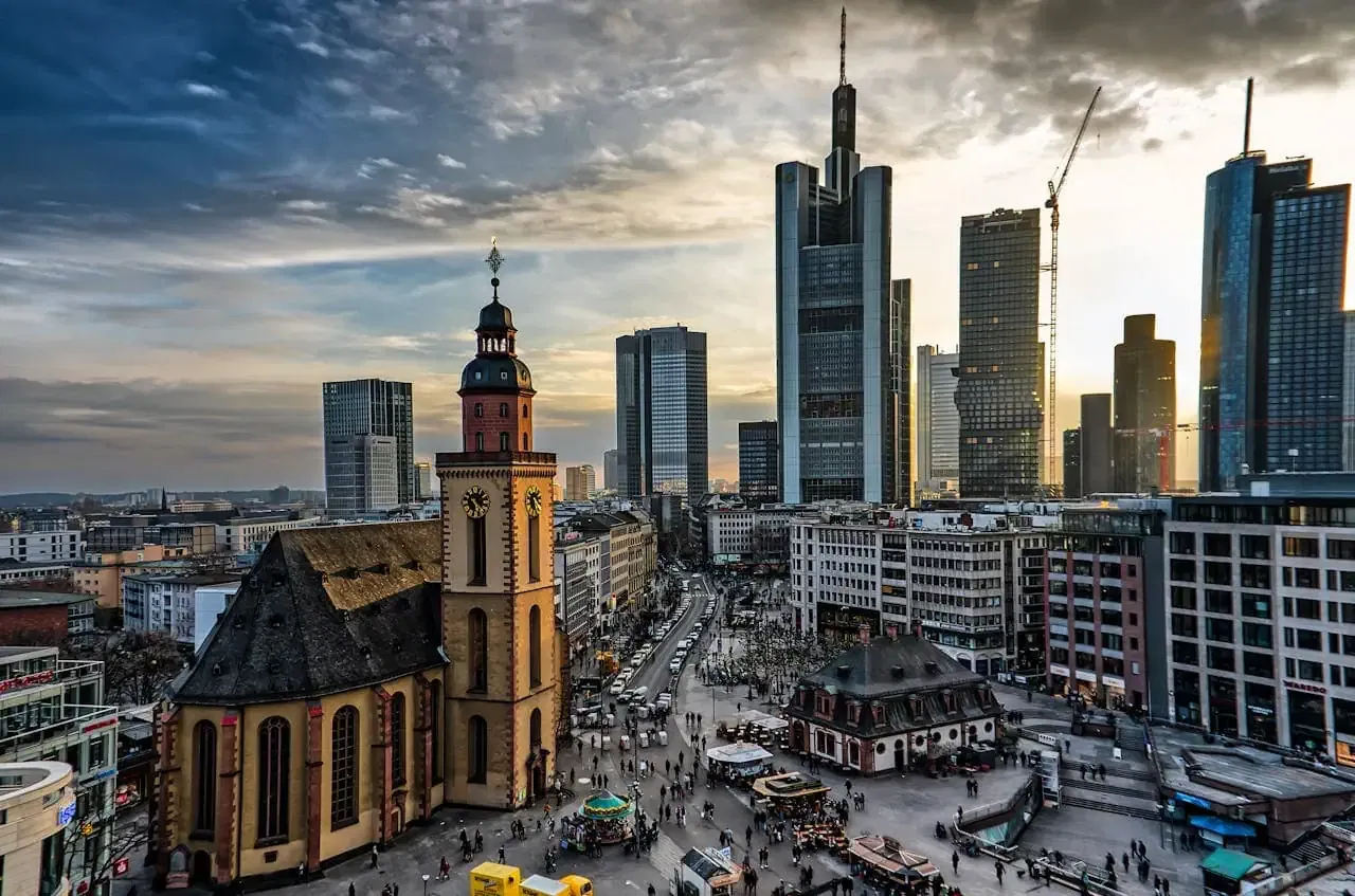 Eine Stadtansicht von Frankfurt bei Sonnenuntergang fängt die Essenz der Stadt ein und verbindet historische und moderne Architektur. Auffällig ist die Katharinenkirche mit ihrem Glockenturm. Wolkenkratzer erheben sich unter einem dramatischen Himmel und blicken auf geschäftige Straßen voller lebhafter Einkaufsmöglichkeiten in Frankfurt.