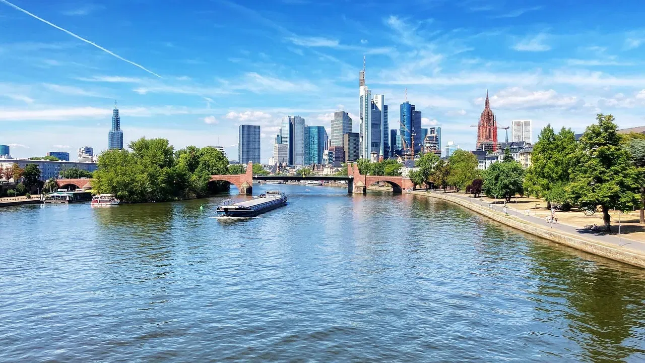 Eine ruhige Flussszene mit einem großen Lastkahn, der auf dem Wasser treibt, flankiert von von Bäumen gesäumten Ufern. Moderne Wolkenkratzer und historische Gebäude ragen vor einem klaren blauen Himmel empor, während im Mittelgrund eine elegante Brücke, die an einen automatisch gespeicherten Entwurf erinnert, den Fluss überspannt.