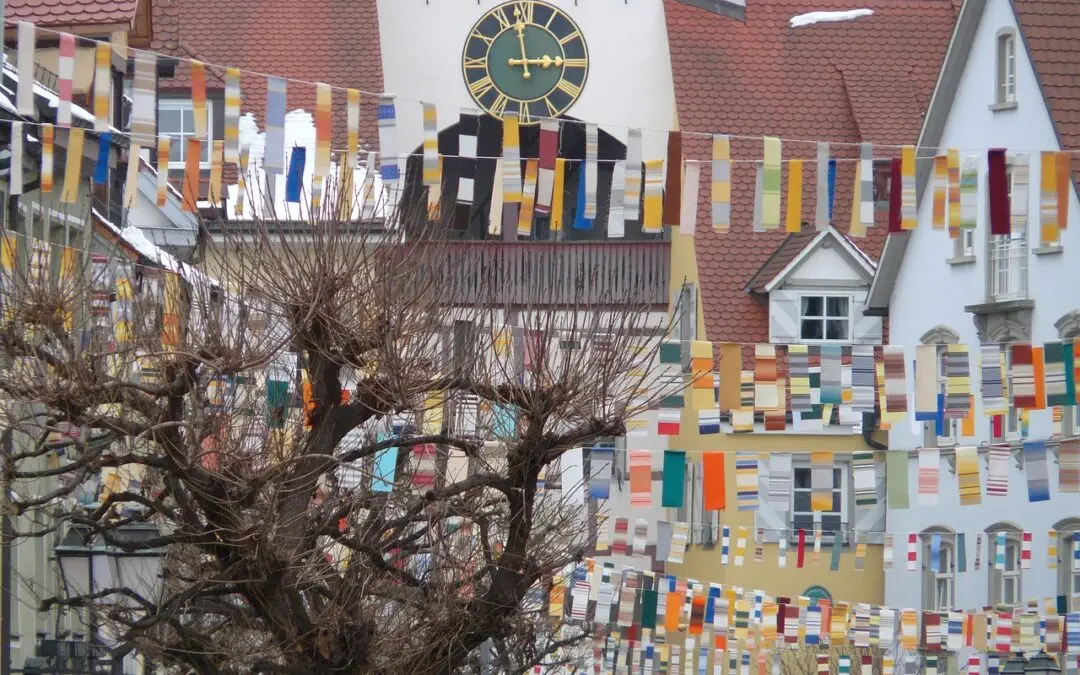 Eine Straße in Frankfurt ist mit bunten, rechteckigen Flaggen geschmückt, die zwischen den Gebäuden hängen. Im Hintergrund ist ein Uhrturm mit einem großen gelb-schwarzen Ziffernblatt zu sehen. Entlang der Straße stehen kahlköpfige Bäume, und Häuser mit roten Ziegeldächern umgeben das Gebiet und deuten auf bevorstehende lokale Ereignisse hin.