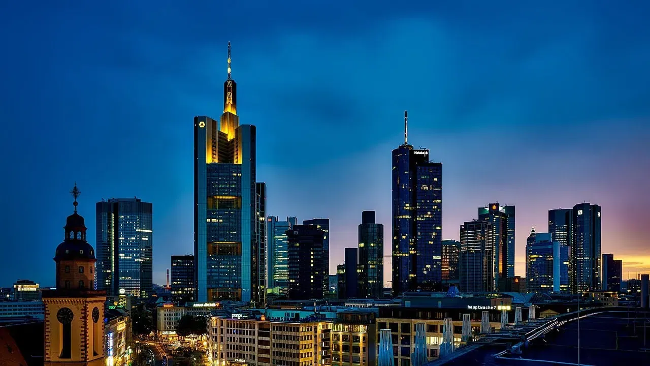 Eine Skyline in der Abenddämmerung mit beleuchteten Wolkenkratzern vor einem blau-violetten Himmel. Im Vordergrund ragt ein historisches Gebäude mit einer Kuppel empor. Das höchste Gebäude, das Frankfurts Finanzszene widerspiegelt, zeichnet sich durch einen markanten, gelb beleuchteten Turm aus.