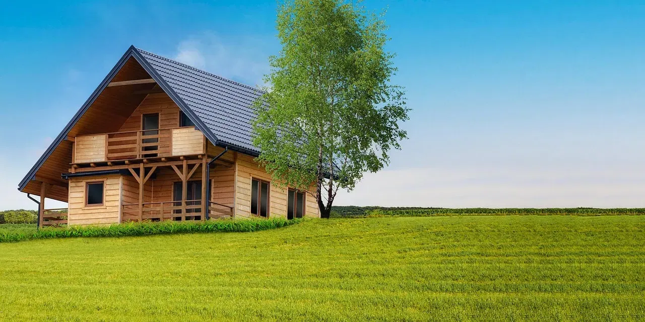 Ein gemütliches Ferienhaus aus Holz mit steilem blauem Dach steht auf einer Wiese unter dem klaren blauen Himmel. Ein hoher Baum neben dem Haus bringt Grün in die ruhige, sonnendurchflutete Landschaft und schafft einen idealen Zufluchtsort für alle, die Tipps und Tricks für Wochenendausflüge suchen.