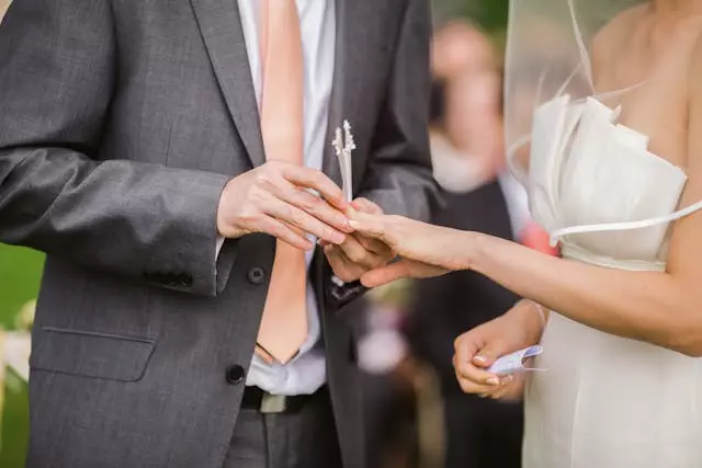 Bei ihrer Hochzeit im Freien steckt ein Bräutigam in grauem Anzug und pfirsichfarbener Krawatte der Braut einen Ring an den Finger. Sie trägt ein weißes Kleid und einen Schleier und verwirklicht damit ihre Träume. Der Hintergrund ist sanft verschwommen, was den Fokus auf ihre Hände und Ringe verstärkt.