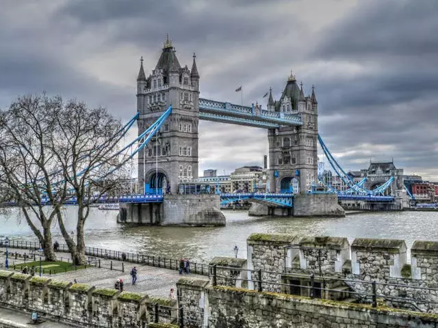 Erleben Sie einen Städtetrip nach London mit einem atemberaubenden Blick auf die Tower Bridge mit ihren ikonischen Zwillingstürmen und Hängeelementen. Unter ihnen fließt die Themse, mit bewölktem Himmel und kahlen Bäumen im Vordergrund, während Menschen am Uferweg entlangschlendern.