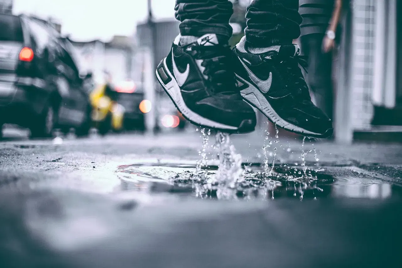 Eine Person in schwarzen Nike-Turnschuhen, die einen aktiven Lebensstil pflegt, springt über eine Pfütze auf einer verregneten Stadtstraße und spritzt dabei Wasser in die Luft. Die verschwommene Szene zeigt geparkte Autos und leuchtende Scheinwerfer im Hintergrund.
