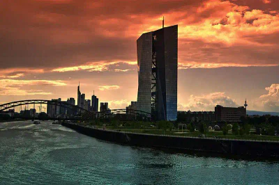 Ein moderner Wolkenkratzer ragt hoch über dem Fluss in einer Stadtlandschaft mit einem orange-roten Sonnenuntergangshimmel empor. Die Szene umfasst die historische EZB-Brücke, mehrere andere Hochhäuser im Hintergrund und Grünflächen entlang des Wassers.